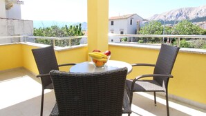 Un balcone con due sedie in vimini, un tavolino rotondo, una ciotola di frutta e una vista su edifici e montagne sullo sfondo.