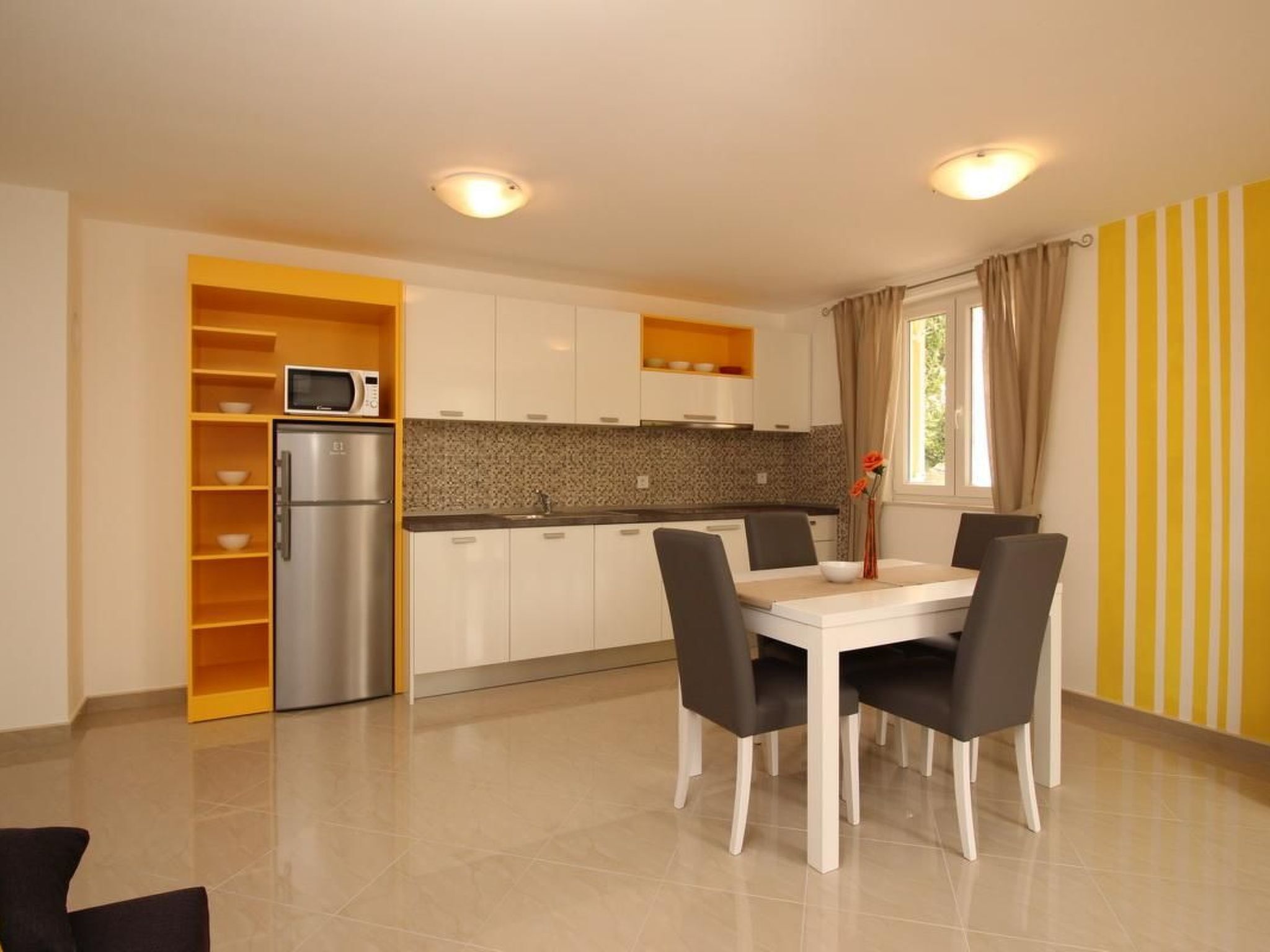 Una cocina moderna con armarios blancos, un refrigerador de acero inoxidable y un microondas. Hay una mesa de comedor con cuatro sillas y un jarrón con flores.