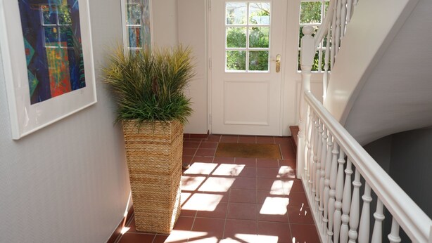 Un couloir avec une porte blanche, un grand pot de fleurs en osier et un escalier avec des balustrades blanches.