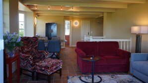 Un salon avec un canapé rouge, un fauteuil bleu et des chaises à motifs. On y trouve une petite table avec une lampe, une grande fenêtre et une porte qui donne sur une autre pièce.