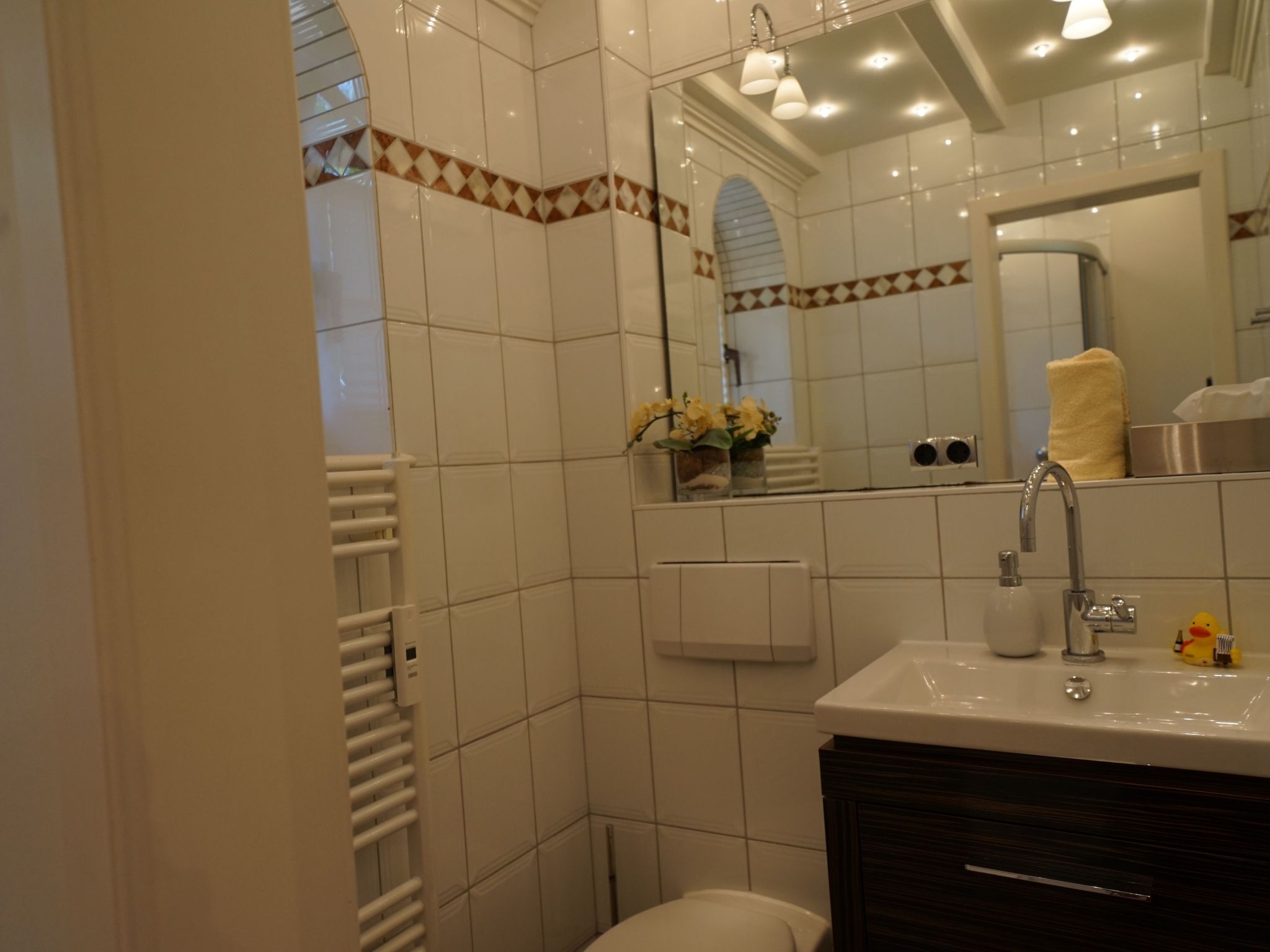 Ein Badezimmer mit weißen Fliesen, einem Waschbecken, einem Spiegel, einem Handtuchhalter und einer Toilette.