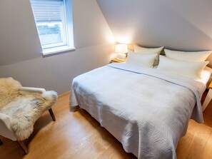 A bedroom with a bed, a nightstand, a lamp, a fur chair, and a window with blinds. - Heefwai - H2W6 - in Morsum by Interhome (Archsum)