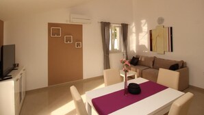 A living room with a sofa, a dining table with chairs, a TV, and a window with curtains. - Gorica II A2+2 br.8 by Interhome (Krk/Baška)