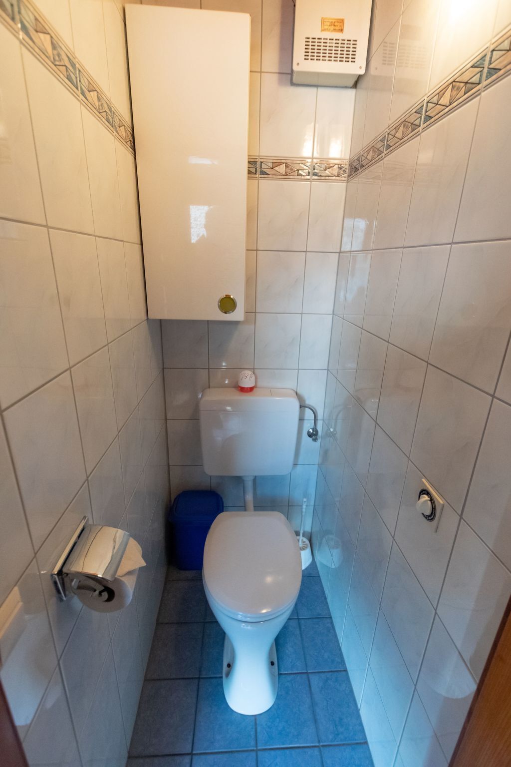 Ein kleines Badezimmer mit Toilette, blauem Mülleimer und weißem Toilettenpapierhalter.