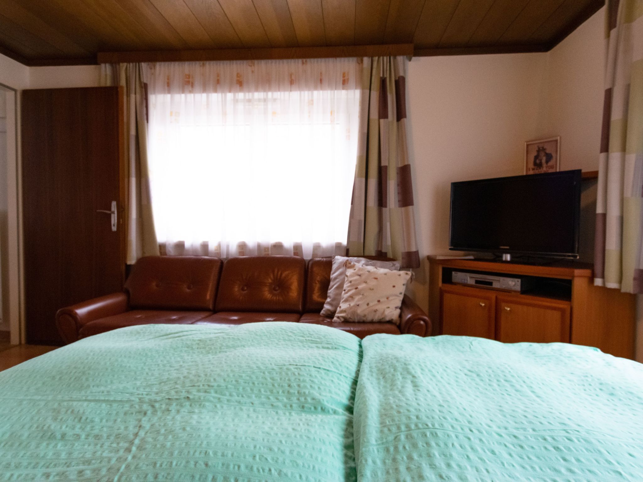 Ein Zimmer mit einem Bett, einem braunen Ledersofa, einem Fernseher auf einer Holzkommode und einem Fenster mit Vorhängen.