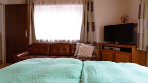 Quarto com cama, sofá de couro marrom, televisão sobre um armário de madeira e uma janela com cortinas.