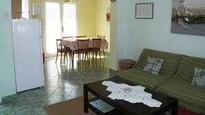 Un salon avec un canapé vert, une table basse ornée d’un chemin de table en dentelle, un coin repas avec une table et des chaises, et une cuisine équipée d’un réfrigérateur.