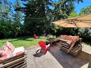 En udendørs patio med et træbord, røde stole, en sofa med mønstrede puder og en stor parasol.
