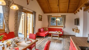 Une pièce chaleureuse avec un plafond en bois, un canapé rouge, une table à manger où sont disposés des aliments pour le petit-déjeuner, et une grande fenêtre avec des rideaux.