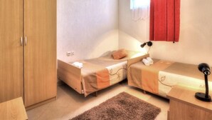 A small, well-lit room with two single beds, a wooden wardrobe, a desk with a lamp, and a window with red curtains. - Grozdana Nr.1A by Interhome (Krk/Baška)