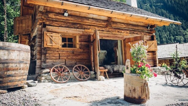 Une cabane en bois rustique, dotée d’un porche en bois, d’une grande roue et d’un pot de fleurs.