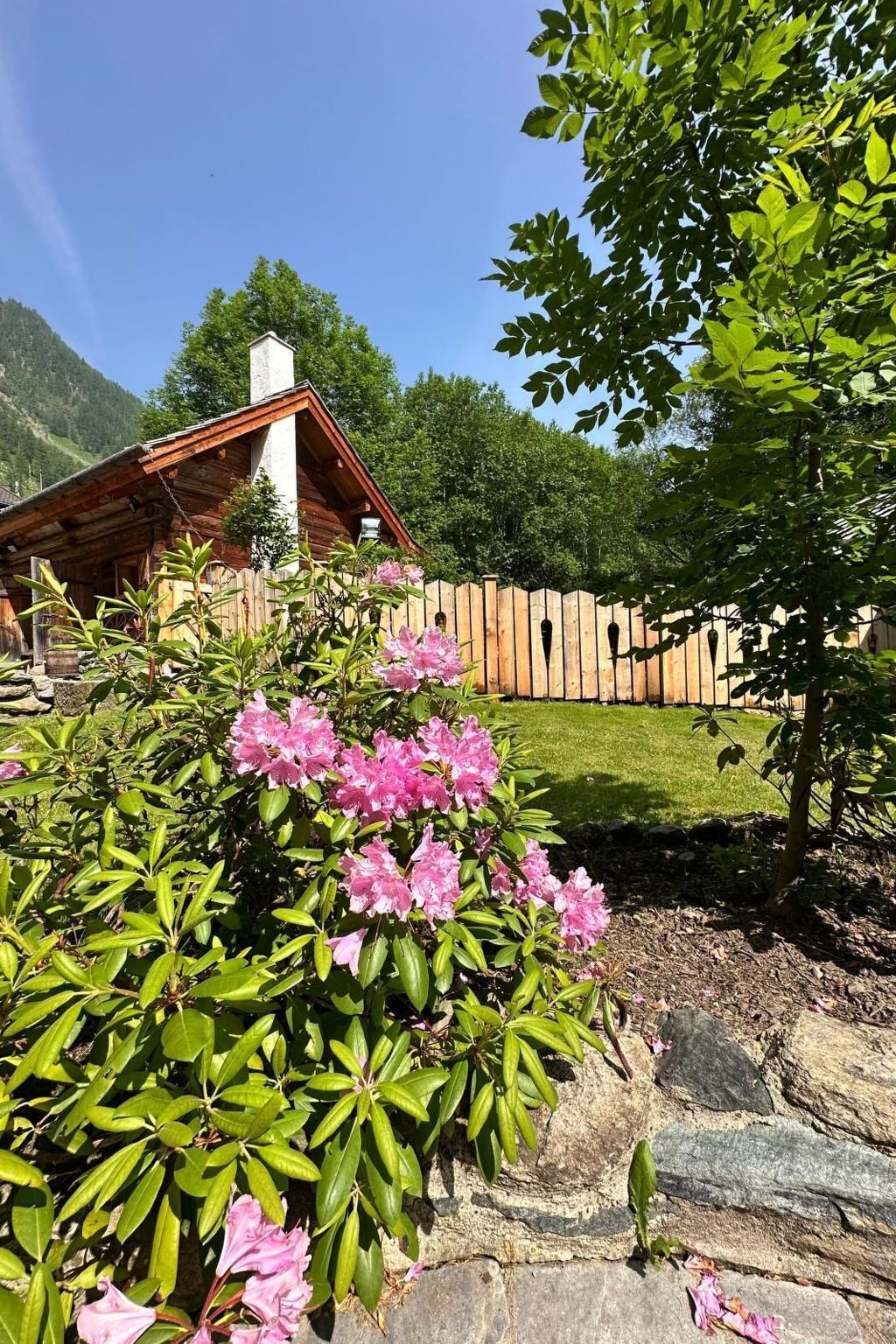 Une cabane en bois, construite sur des fondations en pierre, entourée d’une clôture en bois et bordée d’une pelouse.