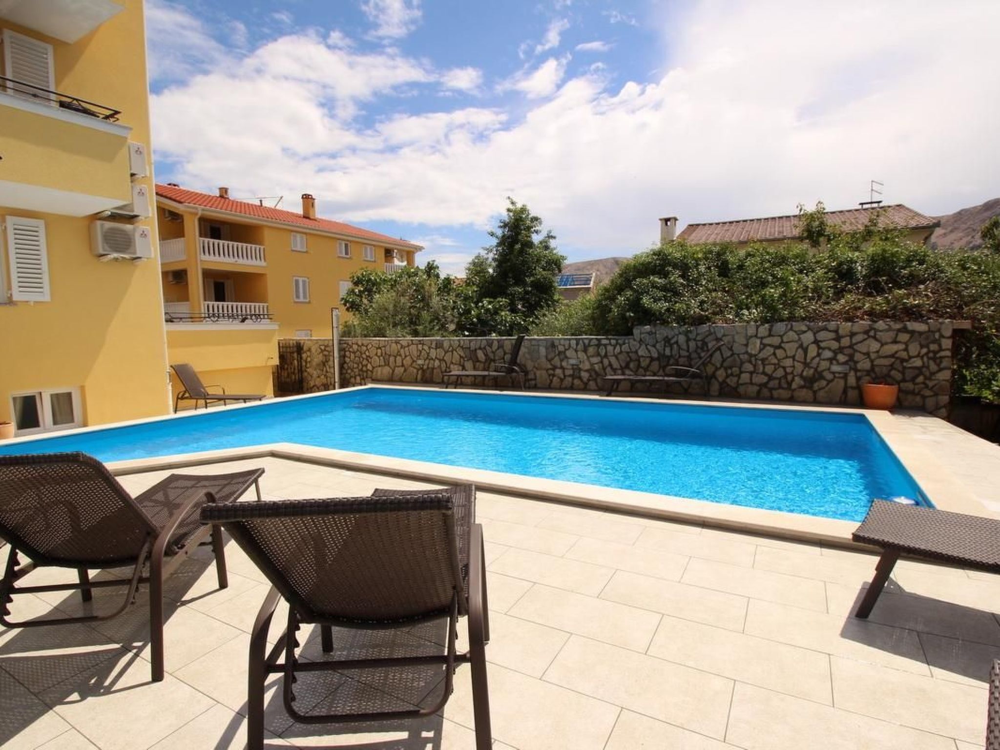 Une piscine avec des chaises longues, un mur en pierre et un bâtiment jaune en arrière-plan.