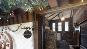 Interior rústico com uma roda de madeira, lenha empilhada e piso coberto por um tapete vermelho.