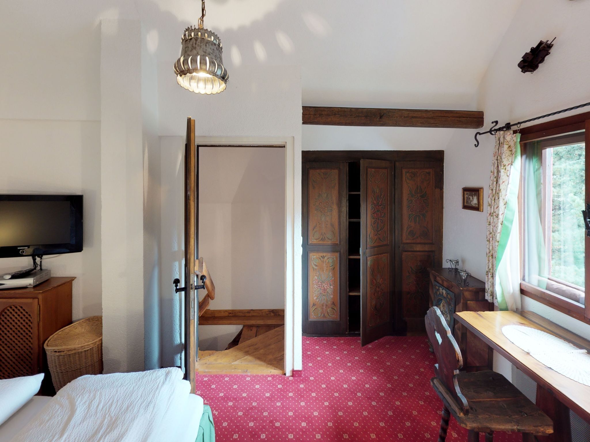 A bedroom with a bed, a television, a wooden desk, a window with curtains, and a staircase leading to another room.