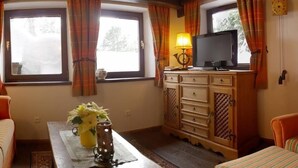 Quarto acolhedor com vigas de madeira, uma televisão sobre um móvel, um vaso com flores e uma janela com cortinas.
