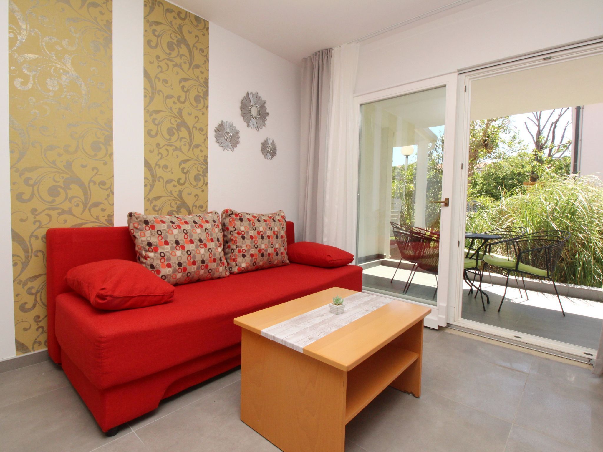 Un canapé rouge agrémenté de coussins à motifs, une table basse en bois et une porte vitrée donnant sur un espace extérieur équipé d’une table et de chaises.