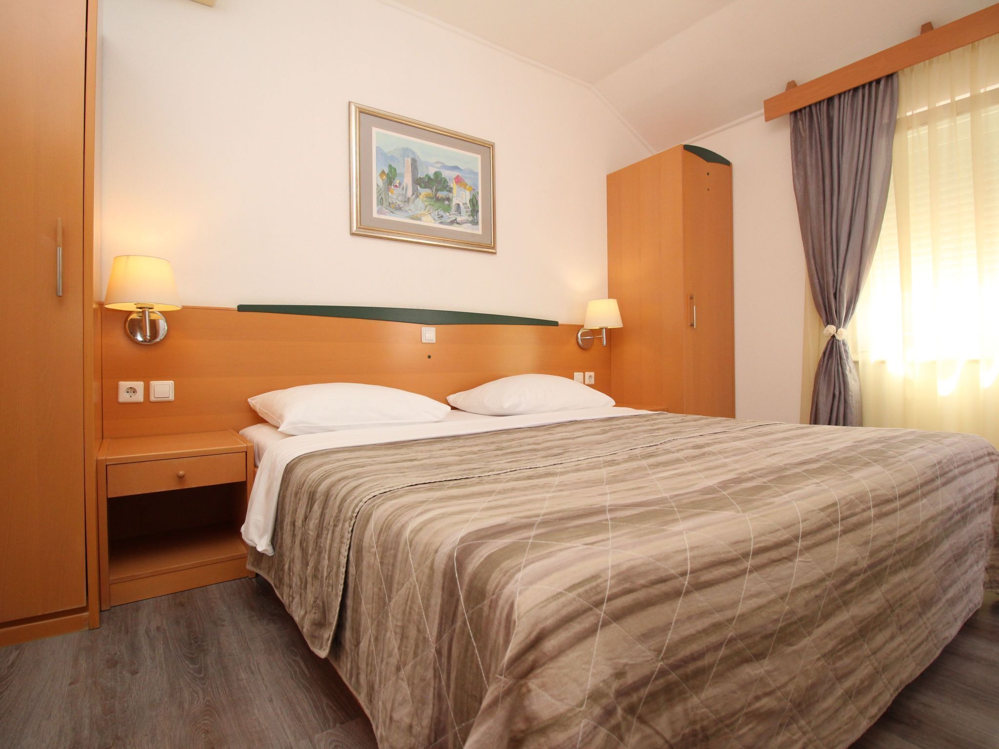A hotel room with a bed, two bedside lamps, a nightstand, a wooden wardrobe, and a painting on the wall.