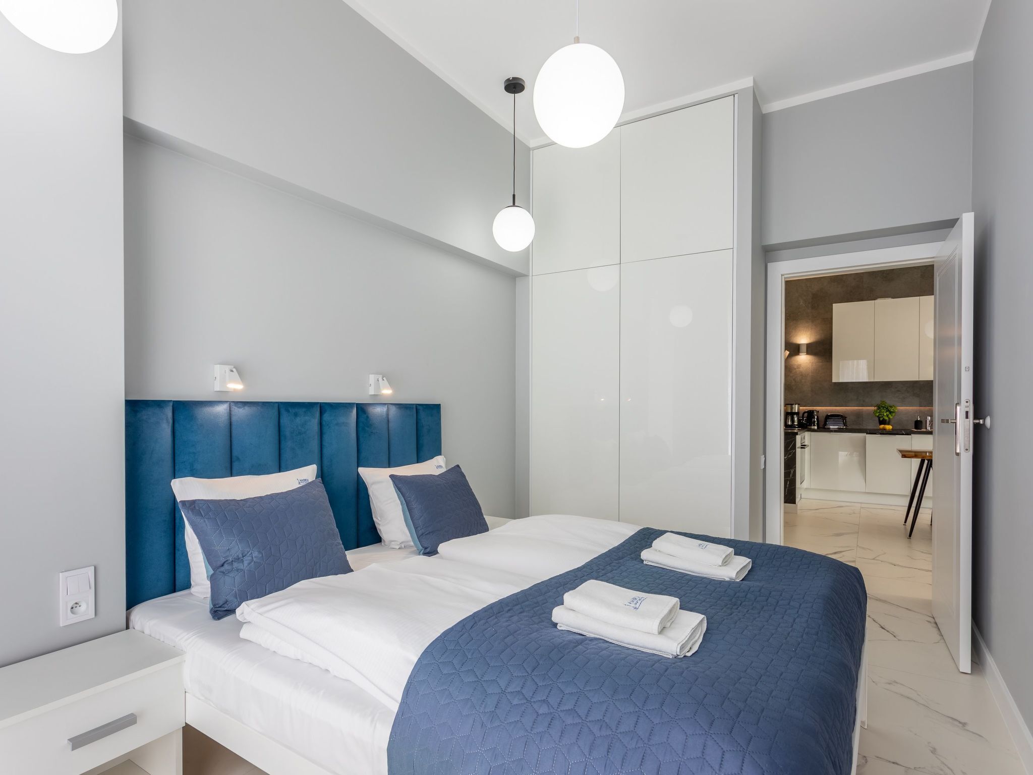 Ein modernes Hotelzimmer mit einem großen Bett, einem blauen Kopfteil und Blick in den Küchenbereich.