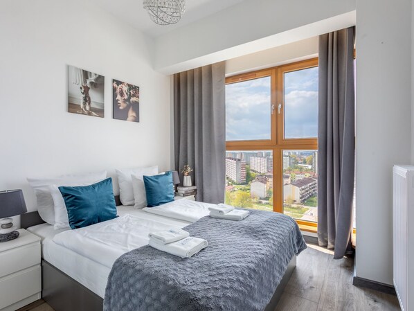 Quarto de hotel moderno com uma cama grande, uma mesa de cabeceira, uma luminária e vista da cidade por uma janela ampla.