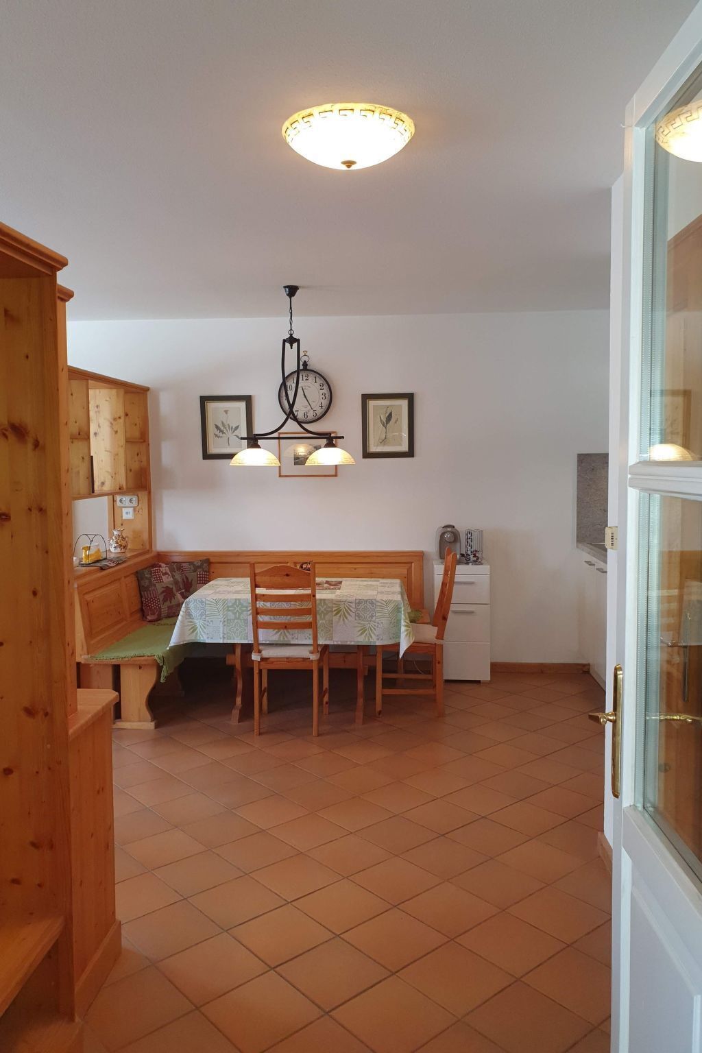 A cozy room with a wooden bench, a table with chairs, a wall clock, and a ceiling light fixture.