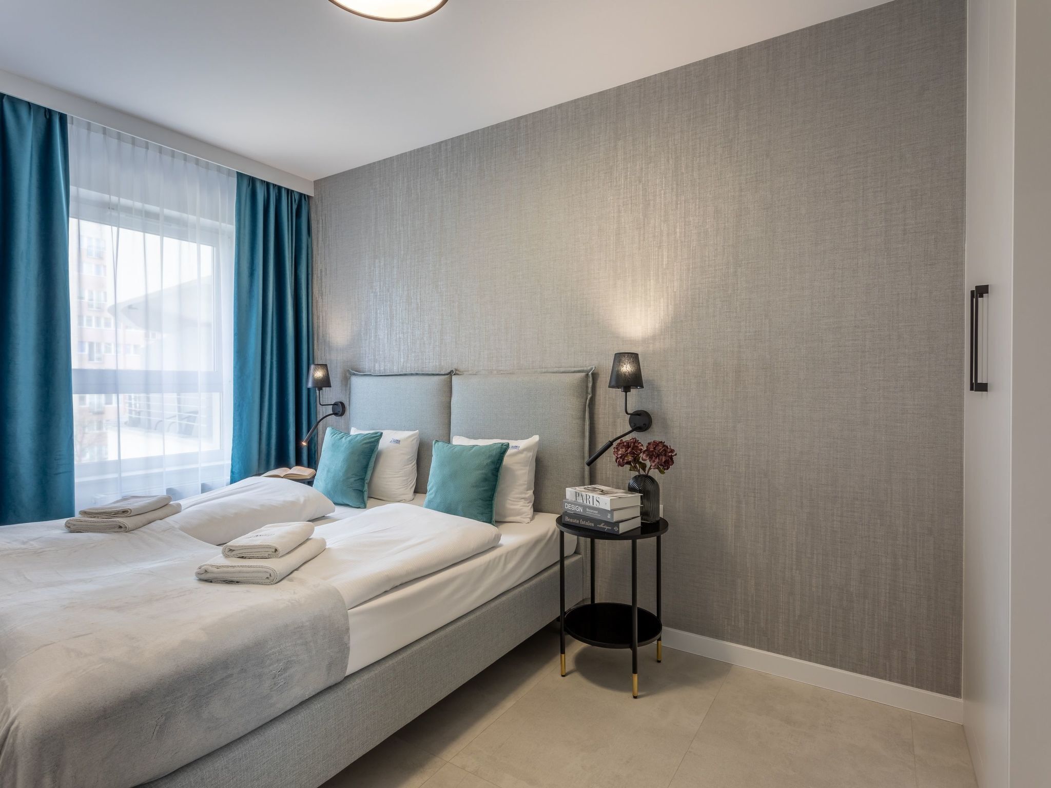 A modern hotel room with a large bed, two bedside lamps, a small side table with a vase of flowers, and a view of the city through sheer curtains.