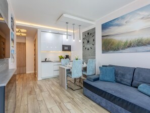 Ett modernt vardagsrum med en soffa, ett matbord och stolar. Det finns ett köksområde med skåp och en mikrovågsugn. En stor klocka hänger på väggen, och en tavla sitter ovanför soffan.
