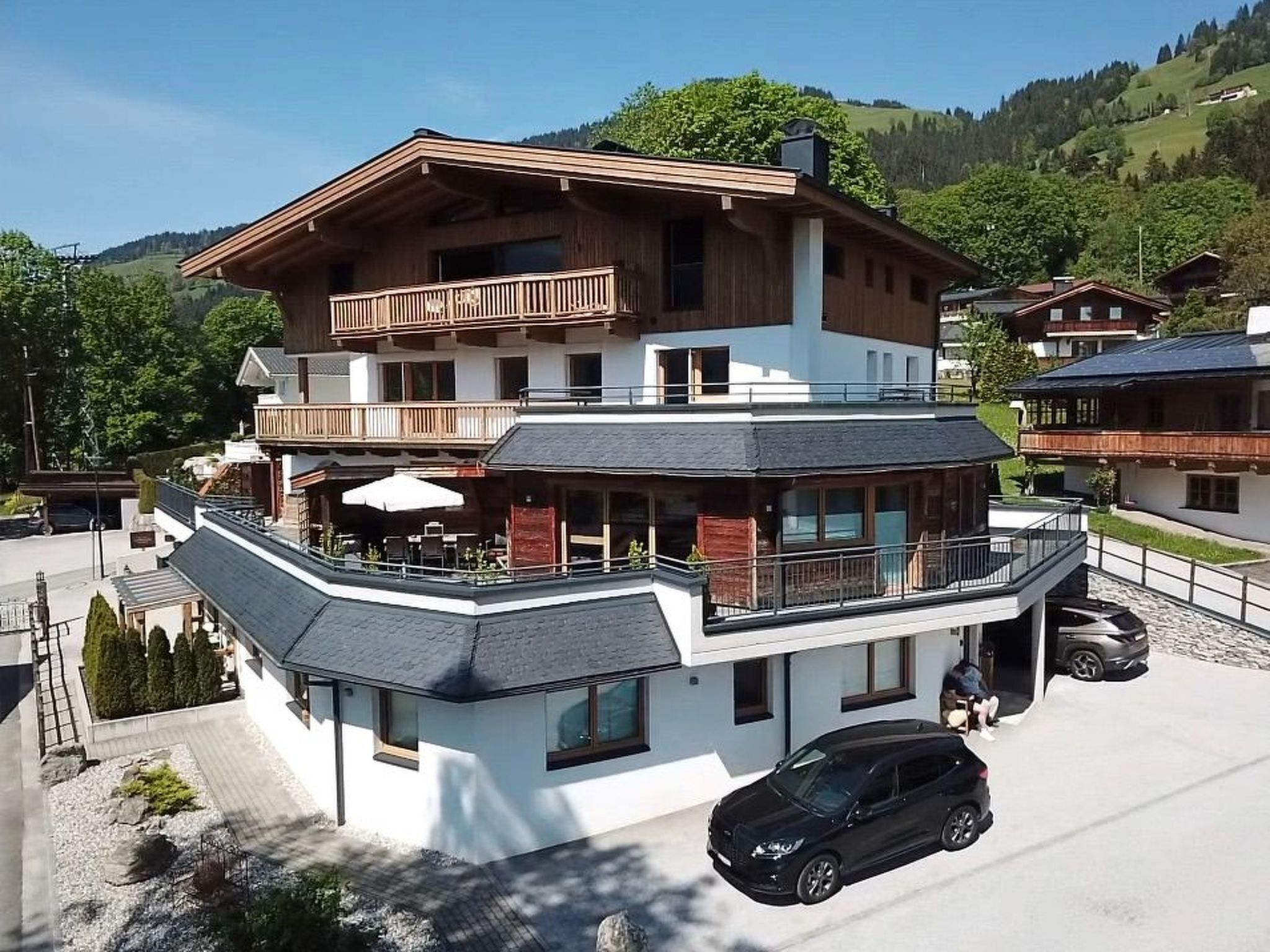A multi-story building with a balcony, a car parked in front, and a mountainous backdrop.