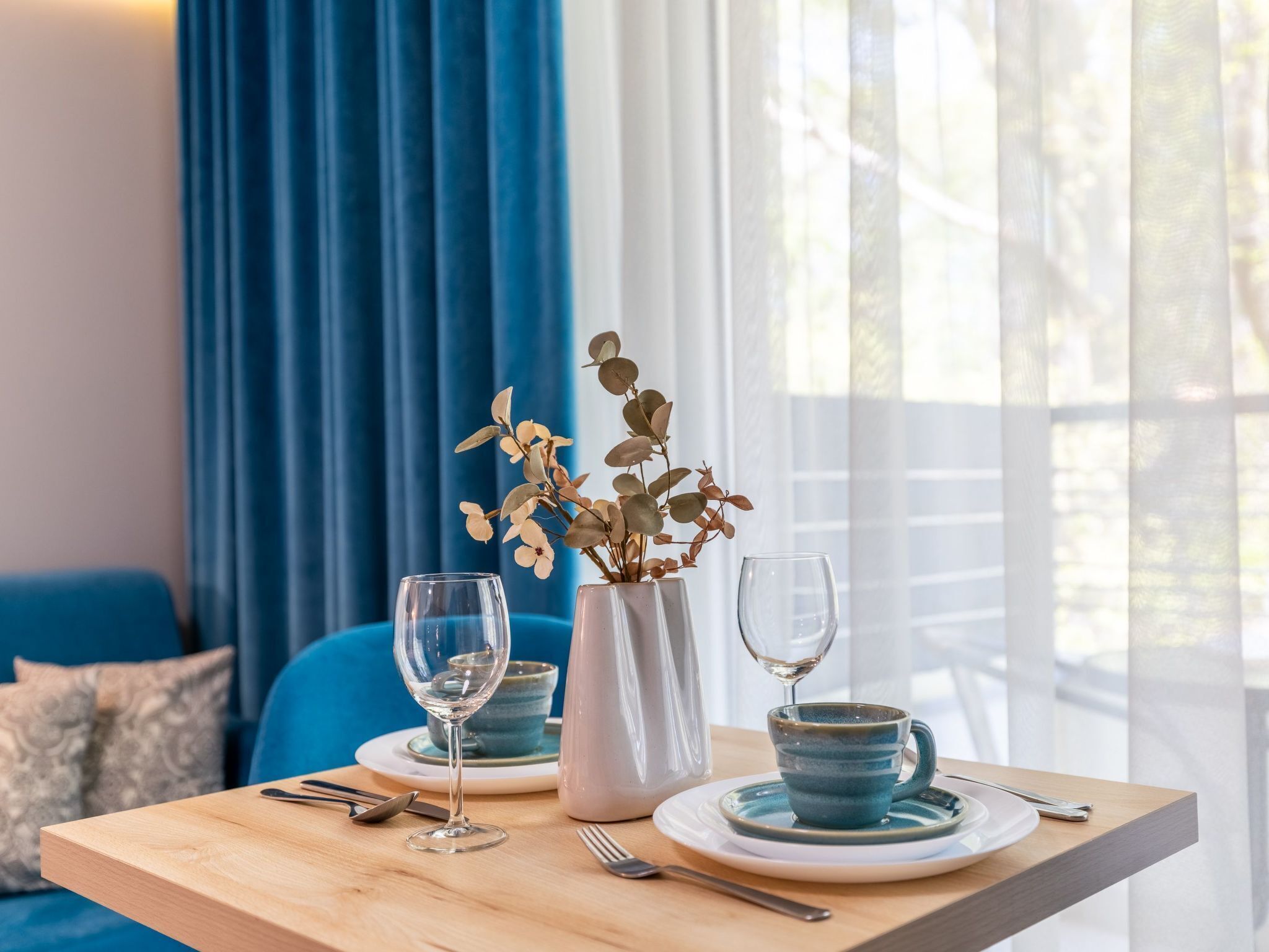 Un tavolo da pranzo con un vaso di fiori secchi, due bicchieri da vino e una tazza di ceramica blu su un tavolo di legno.