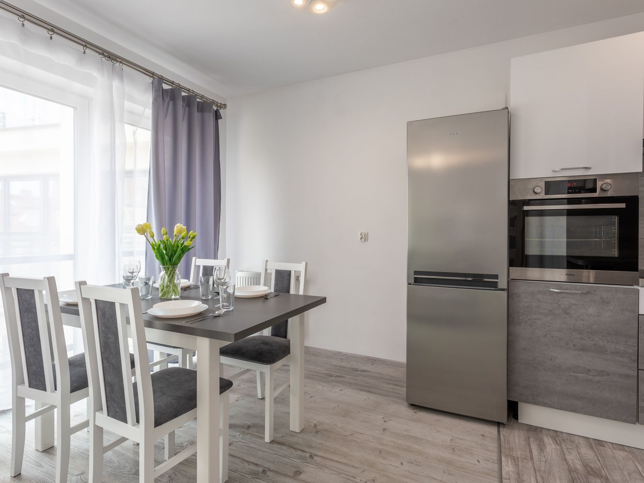 A modern kitchen with a stainless steel refrigerator and oven, a dining table with chairs, and a window with curtains.