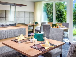 A hotel dining area with a large table set for two, surrounded by patterned chairs, a green upholstered bench, and a view of greenery through large windows. - Wohnung Biohaus in Neukirchen am Großvenediger by Interhome (Neukirchen am Großvenediger)