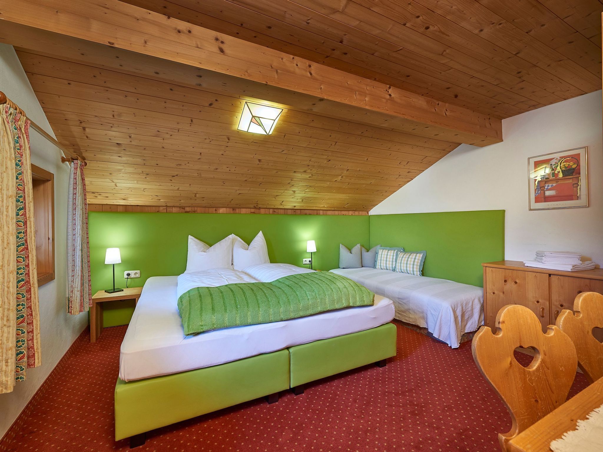 A room with a sloped ceiling, wooden beams, a green headboard, two beds, a nightstand with a lamp, a wooden dresser, and a red carpet.