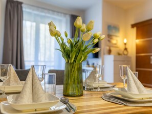 Ein Esstisch ist gedeckt mit weißen Tellern, Servietten und Gläsern, dazu ein Strauß weiße Tulpen in einer Vase und eine Holztischplatte.