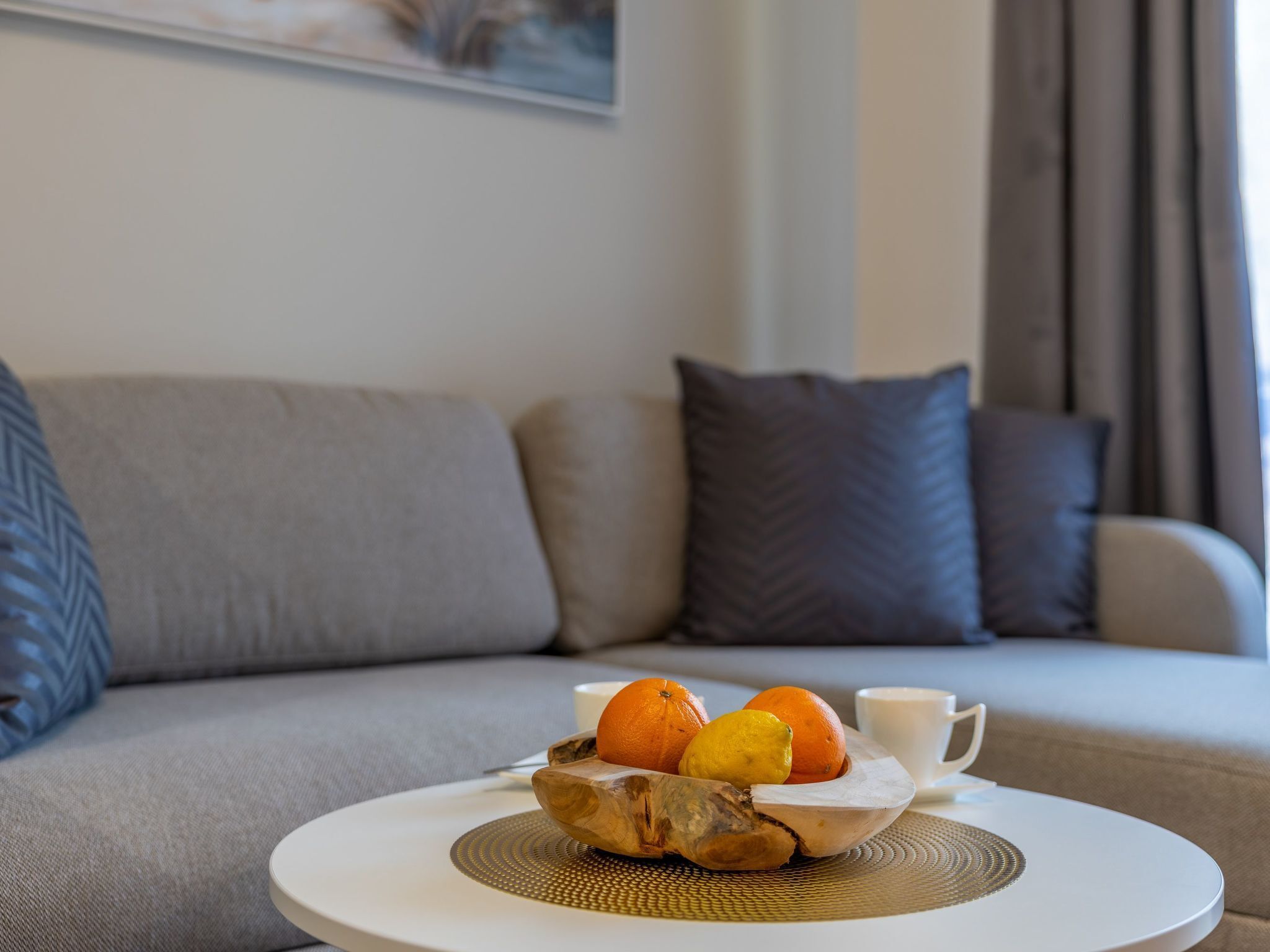 A round white table with a woven placemat, a wooden bowl containing oranges and a lemon, and a white cup. The table is in a living room with a grey sofa and patterned cushions.