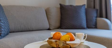 A round white table with a woven placemat, a wooden bowl containing oranges and a lemon, and a white cup. The table is in a living room with a grey sofa and patterned cushions.