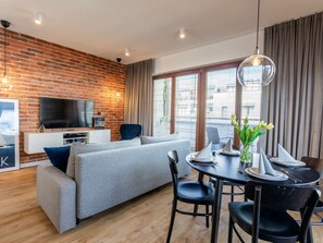 Ein modernes Wohnzimmer mit Backsteinwand, grauem Sofa, Esstisch mit Stühlen und einem an der Wand befestigten Fernseher.