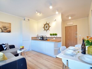 A modern kitchen with white cabinets, a wooden countertop, and a ship's wheel decoration. There is a dining area with a table set for two, a sofa, and a small side table with a plant and a book. - Sand Rose am Yachthafen by Interhome (Swinoujscie)