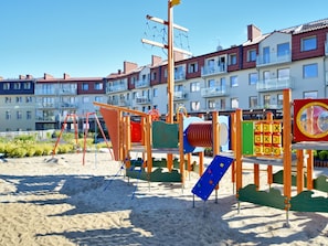 A playground with various play structures, including slides and climbing areas, situated on a sandy area with residential buildings in the background. - Sand Rose am Yachthafen by Interhome (Swinoujscie)