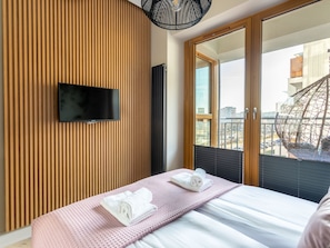 A modern hotel room with a large bed, a flat-screen TV mounted on a wooden panel wall, and a view of the city through large windows. - Tower 26 - Ihr Ferienappartement in Swinemünde by Interhome (Swinoujscie)