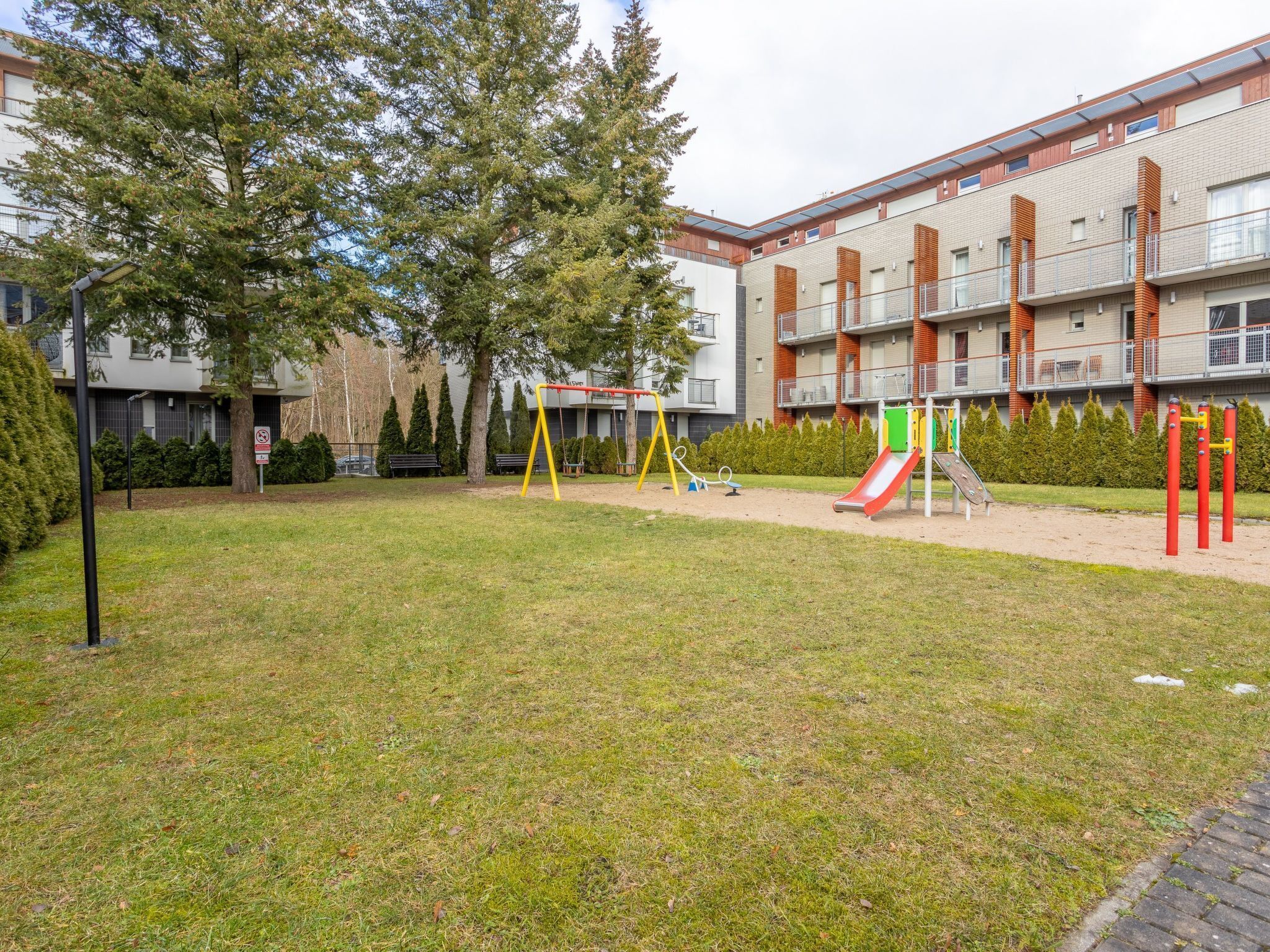 Ein Spielplatz mit Schaukeln und einer Rutsche, umgeben von einer Rasenfläche und Bäumen, mit einem mehrstöckigen Gebäude im Hintergrund.