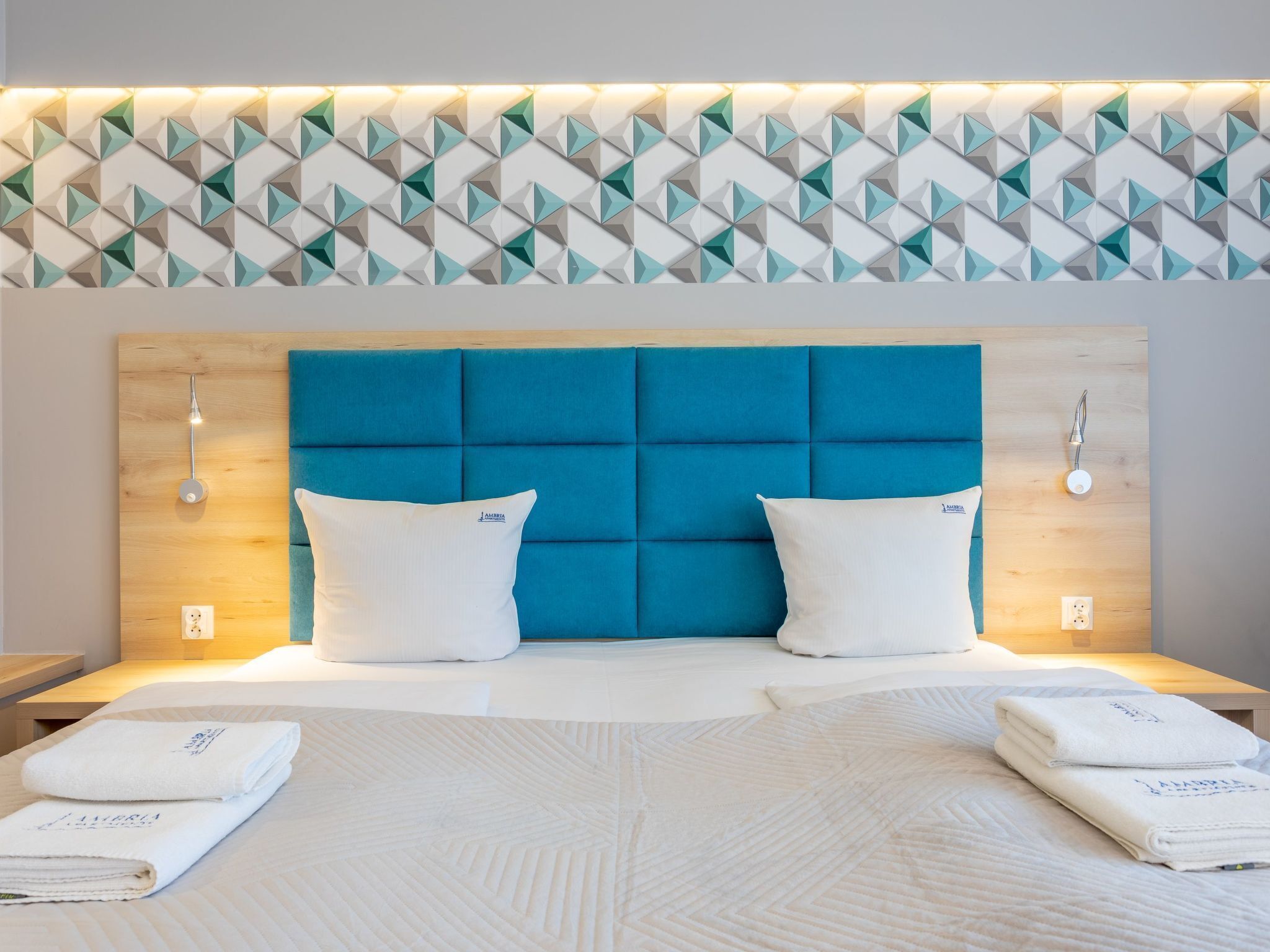 Habitación de hotel con diseño moderno, destacando una pared con motivo geométrico, un cabecero azul con botonadura y toallas ordenadamente dobladas sobre la cama.