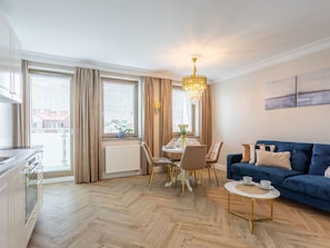 A modern living room with a blue sofa, a dining table with chairs, a chandelier, and large windows with curtains. - Rose 1 in Swinemünde am Meer by Interhome (Swinoujscie)