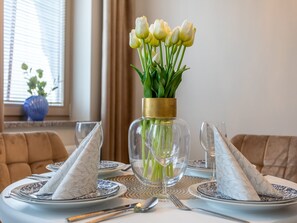 Une table à manger dressée avec de l’argenterie blanche, des serviettes à motifs bleus et blancs, et un vase transparent contenant des tiges vertes et des fleurs blanches.