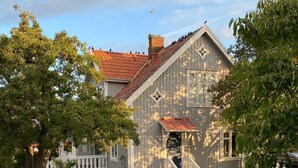 Een huis met een stenen pad dat leidt naar een houten terras, omringd door bomen en een witte hekken.