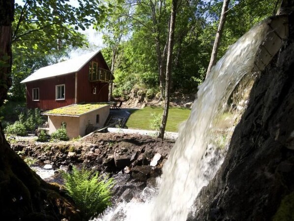 Een rustieke houten hut met een kleine waterval ervoor, omringd door weelderig groen.