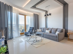 A modern living room with a grey sofa, a black chair, and a glass coffee table. There is a large window with sheer curtains, a view of the outdoors, and a hanging light fixture. - Helle Ferienwohnung Tower 76 auf Usedom by Interhome (Swinoujscie)