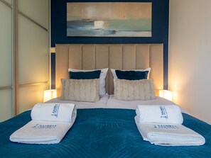 En snyggt bäddad säng med två kuddar, ett blått överkast och två hopvikta handdukar med hotellets logotyp.