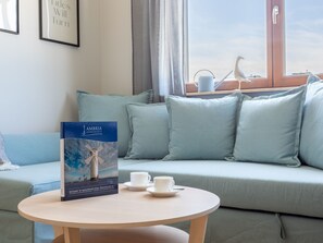 Ein modernes Wohnzimmer mit einem hellblauen Sofa, einem runden Holztisch mit einem Buch und zwei Tassen, sowie einem Fenster mit Blick auf das Meer und einen Vogel.