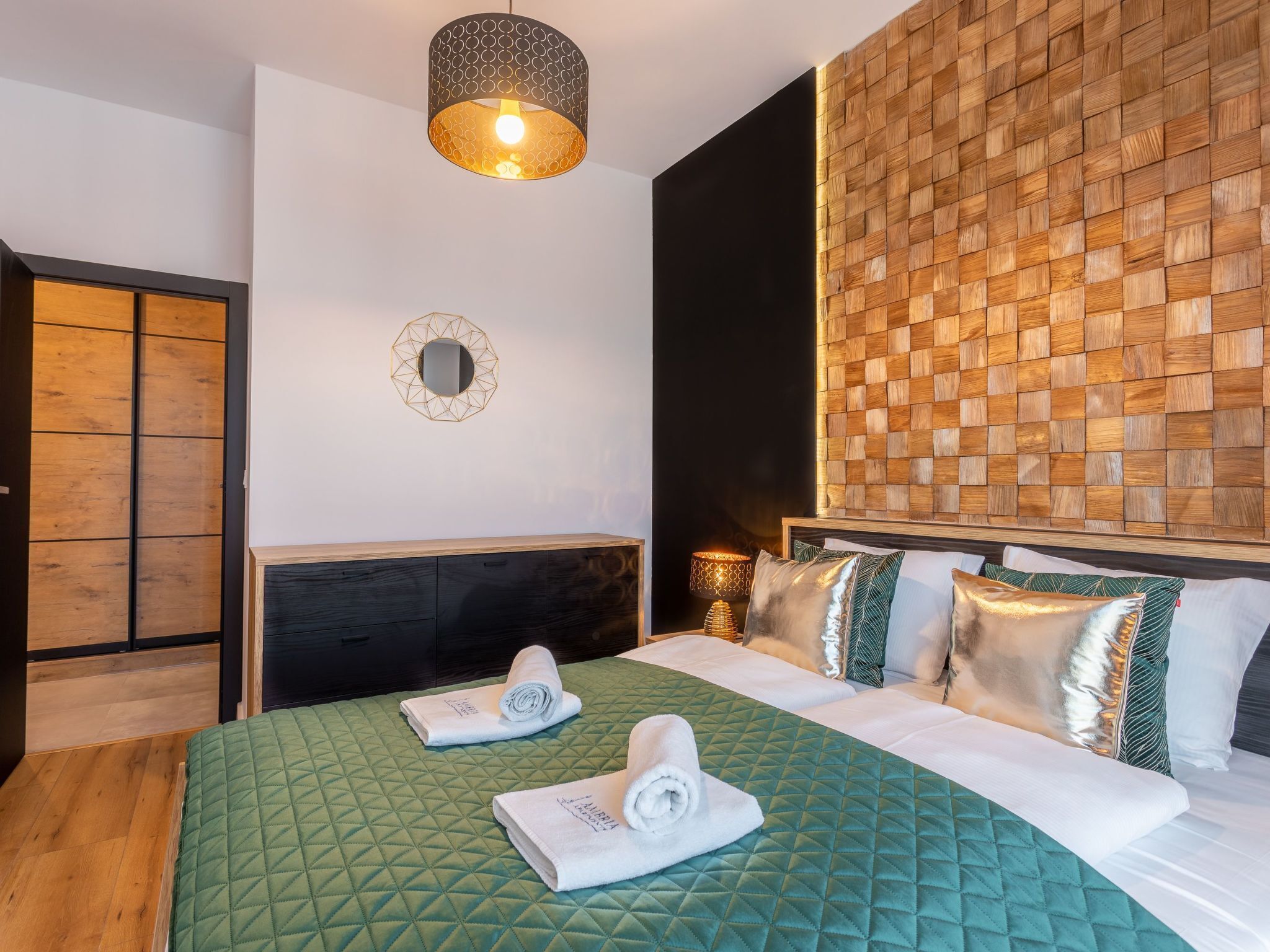 Ein Hotelzimmer mit einer grün gesteppten Bettdecke, einem Holz-Kopfteil, einem geometrischen Spiegel und einer gewebten Wandverkleidung.