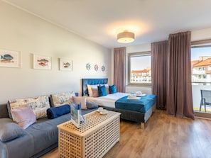 A modern hotel room with a bed, sofa, and a wooden coffee table. There are framed pictures on the wall and a view of buildings outside the window. - Ferienappartement Blue Rose direkt am Park by Interhome (Swinoujscie)